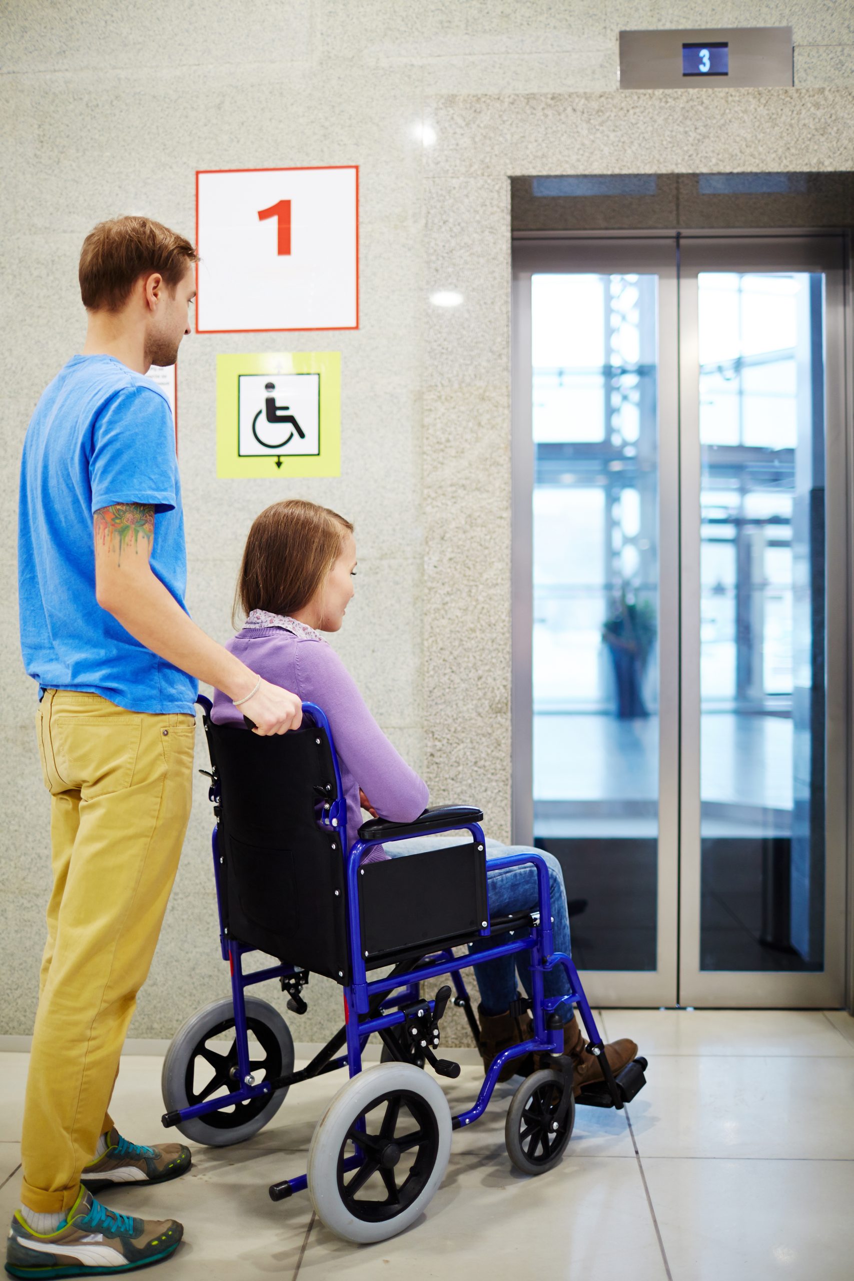 Physically impaired young woman in wheelchair and her assistant waiting for elevator in shopping mall